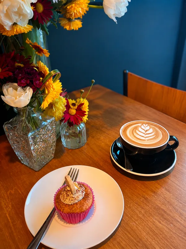 A coffee in a black cup beside a decorated cupcake and flowers